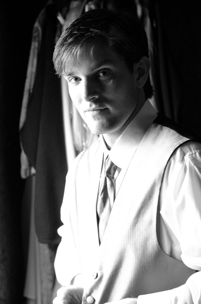 Black-and-white window-light portrait of the groom in shirtsleeves and a patterned vest.