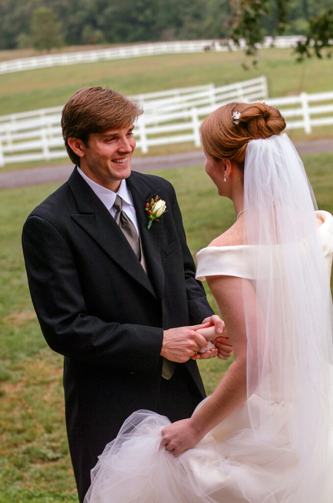 The couple’s first look in a pasture, the groom holding the bride’s hands by a white farm fence.
