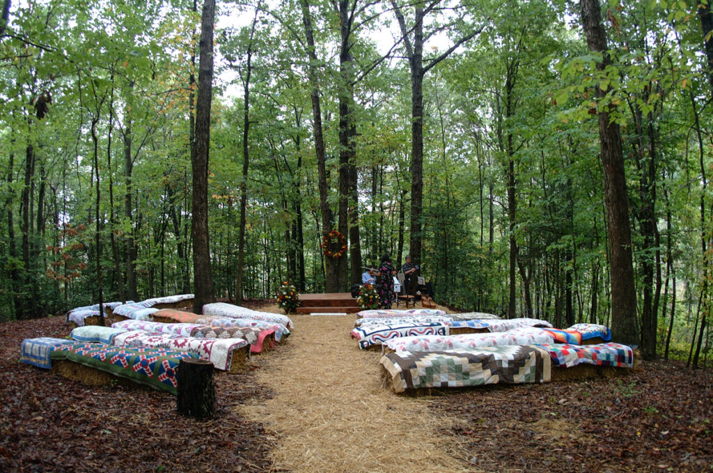 Woodland ceremony seating: hay bales draped in patchwork family quilts along a pine-needle aisle.