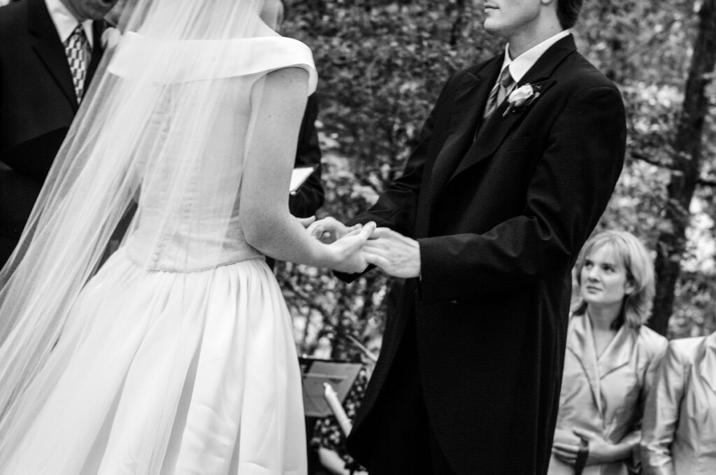 Black-and-white close-up of the couple’s hands during the ring exchange, a guest watching behind.