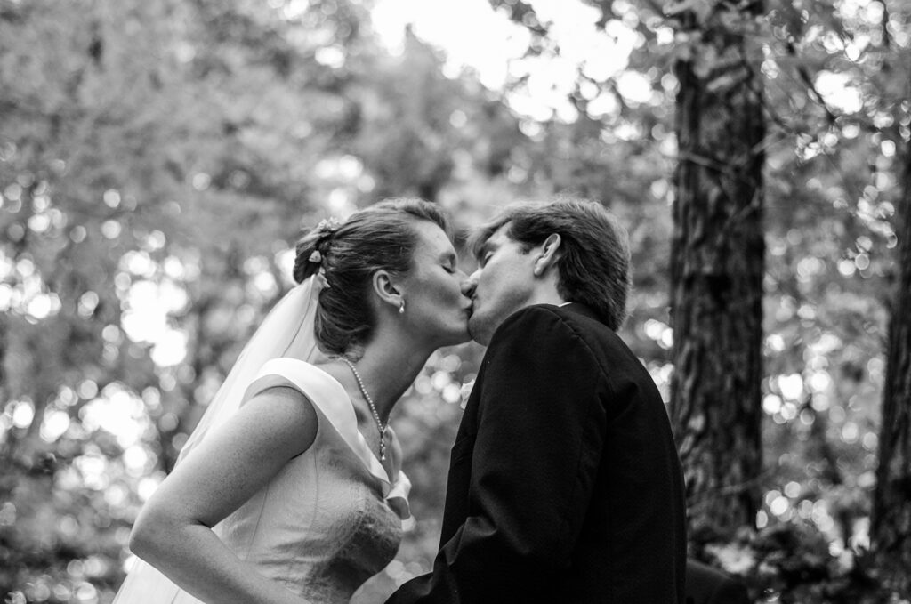 The couple’s first kiss in black and white, dappled woodland light behind them.