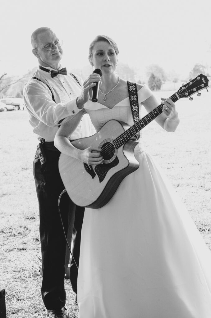 The bride sings and plays guitar at her reception while her father holds the microphone.